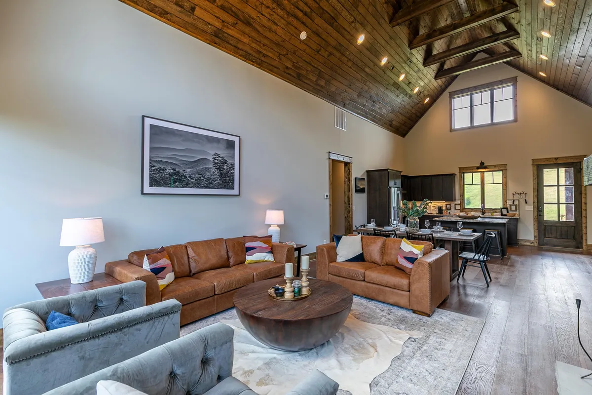 A spacious living area features leather sofas arranged around a round coffee table, with eye-catching artwork on the wall and the kitchen visible in the background. The layout feels open and welcoming, perfect for socializing and group activities.