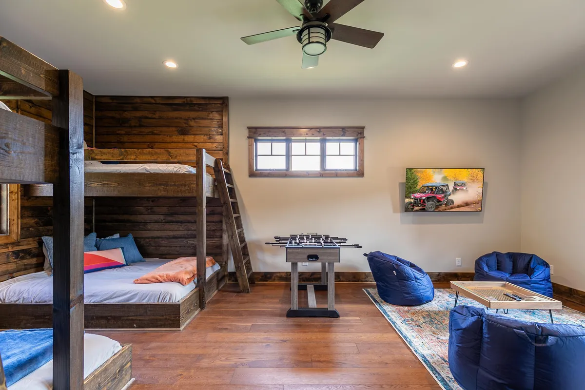 A playful and colorful bunk room featuring sturdy bunk beds, a foosball table at the center, and bean bags for lounging. Framed artwork adds a whimsical touch, and the space is bright with natural light, creating a fun environment for children or group activities.