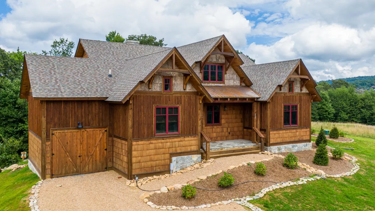 This exterior shot captures the inviting wooden facade of the cabin, accented by red window frames, a landscaped garden, and a stone pathway leading to the porch. The porch is clearly suited for socializing and taking in the surrounding natural beauty, emphasizing the home’s connection to the outdoors.