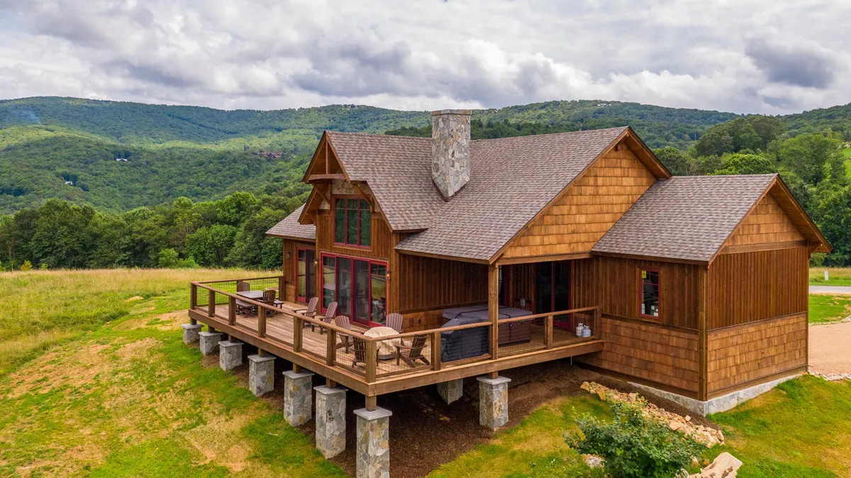 The image captures the cabin's expansive wooden deck framed by sturdy stone pillars and large windows, set against a breathtaking backdrop of rolling Blue Ridge Mountains. The tranquil scene is bathed in sunset light, highlighting inviting spaces for relaxing, dining, and taking in panoramic mountain views.
