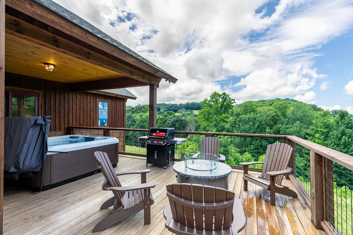 This exterior image presents a cozy outdoor deck featuring a private hot tub, a grill, and adirondack chairs overlooking a scenic mountain view. The space is clearly designed for relaxation, socializing, and outdoor enjoyment, with a tranquil, natural backdrop that evokes the feeling of being immersed in the mountains.