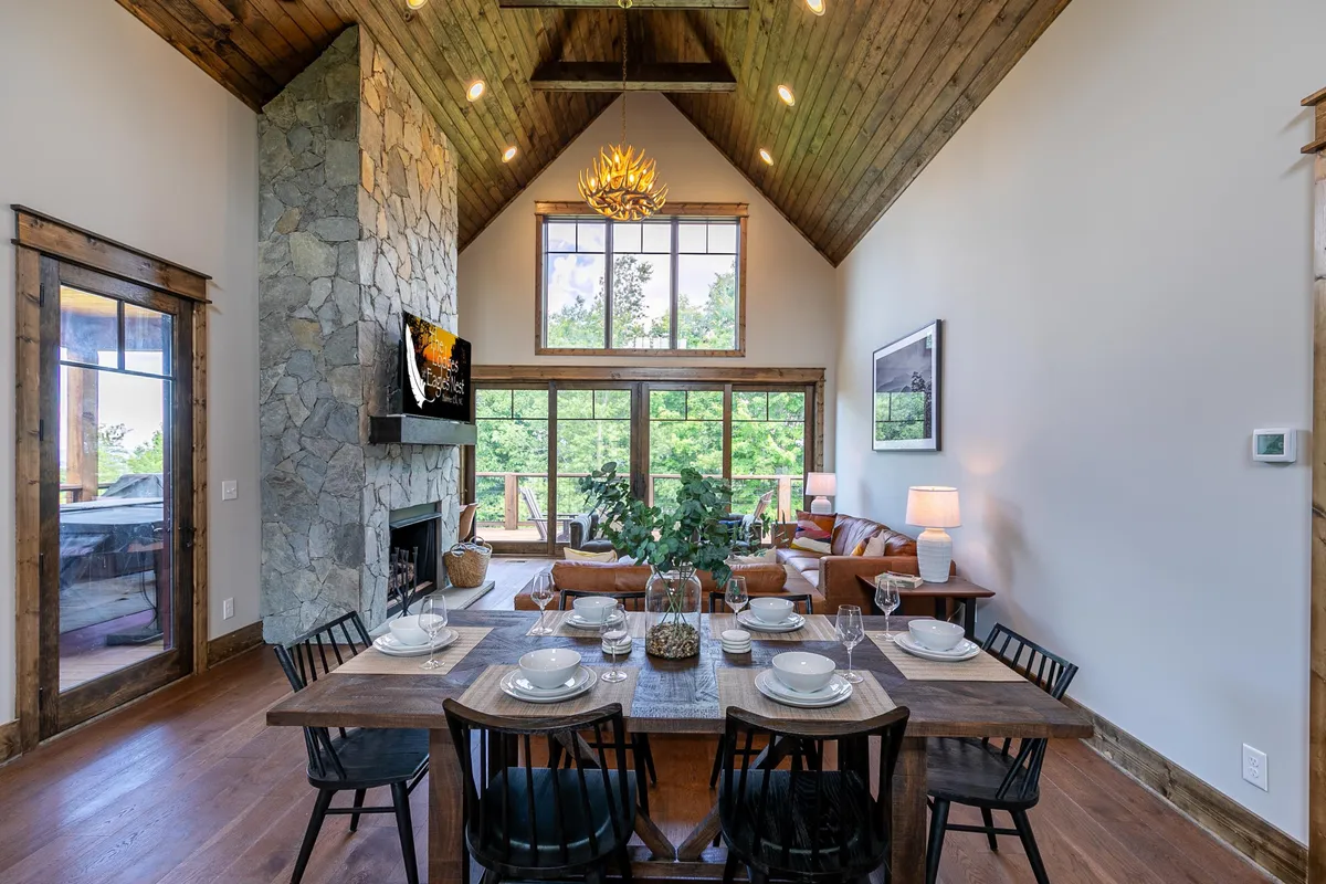 This interior image showcases a spacious open-plan living and dining area with a large dining table set for a meal, a prominent stone fireplace, plush comfortable seating, and expansive windows revealing lush greenery outside. The scene radiates warmth and togetherness, with ample room for family gatherings and communal activities.