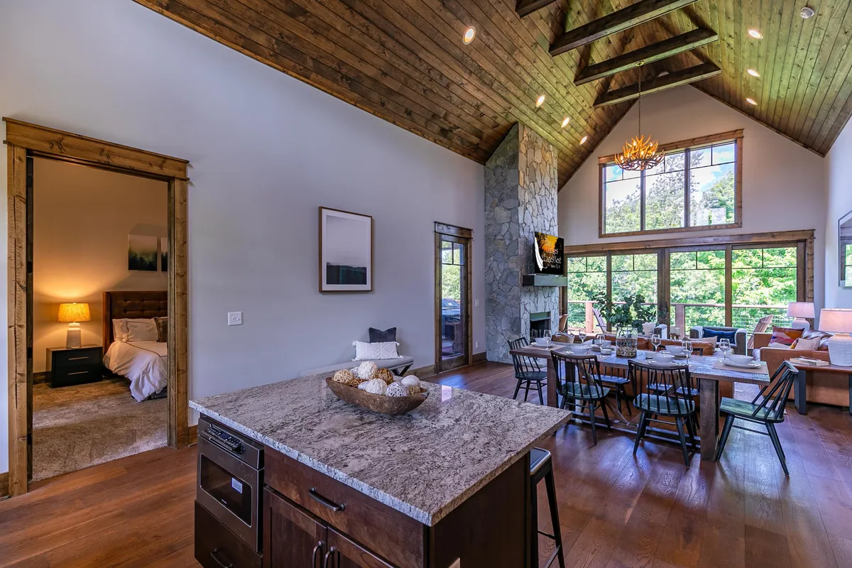 This image captures a spacious interior with a large dining table ready for a meal, a kitchen island adorned with a decorative bowl, big windows letting in ample light and views of greenery, and a stone fireplace anchoring the space. The layout encourages interaction and shared experiences.