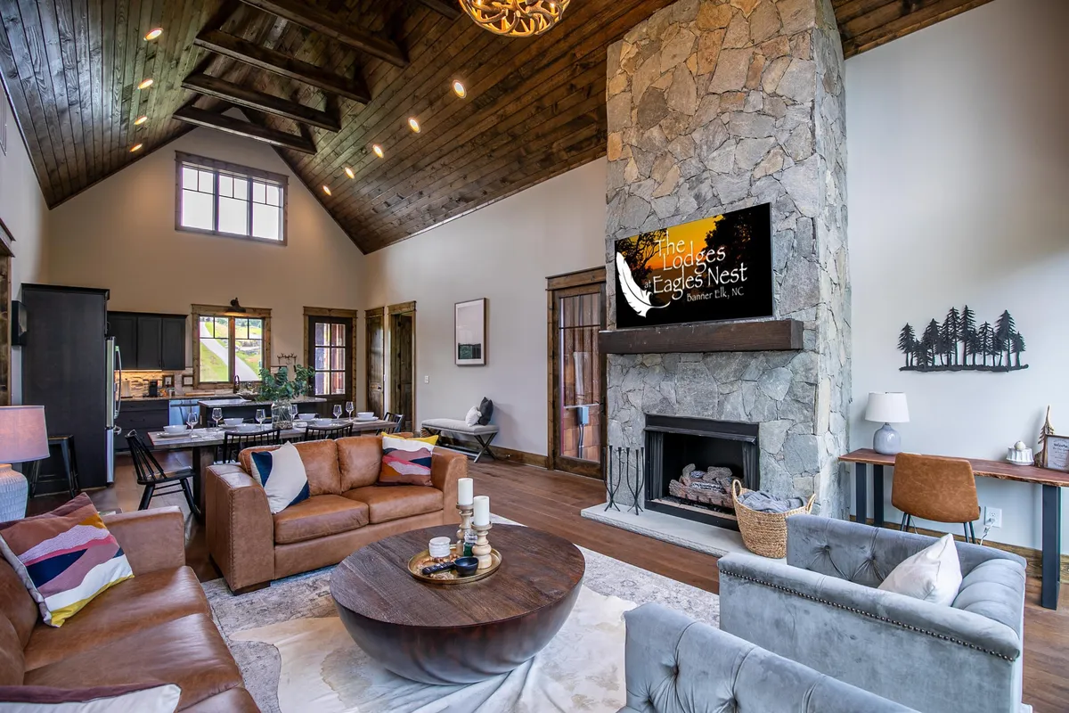 The image depicts a welcoming living space with a stone fireplace, large windows with views, comfortable seating, and a lofty wooden ceiling. The ambiance is cozy and inviting—ideal for snuggling up together by the fire, sharing a glass of wine, or simply enjoying the mountain atmosphere from the comfort of the suite.