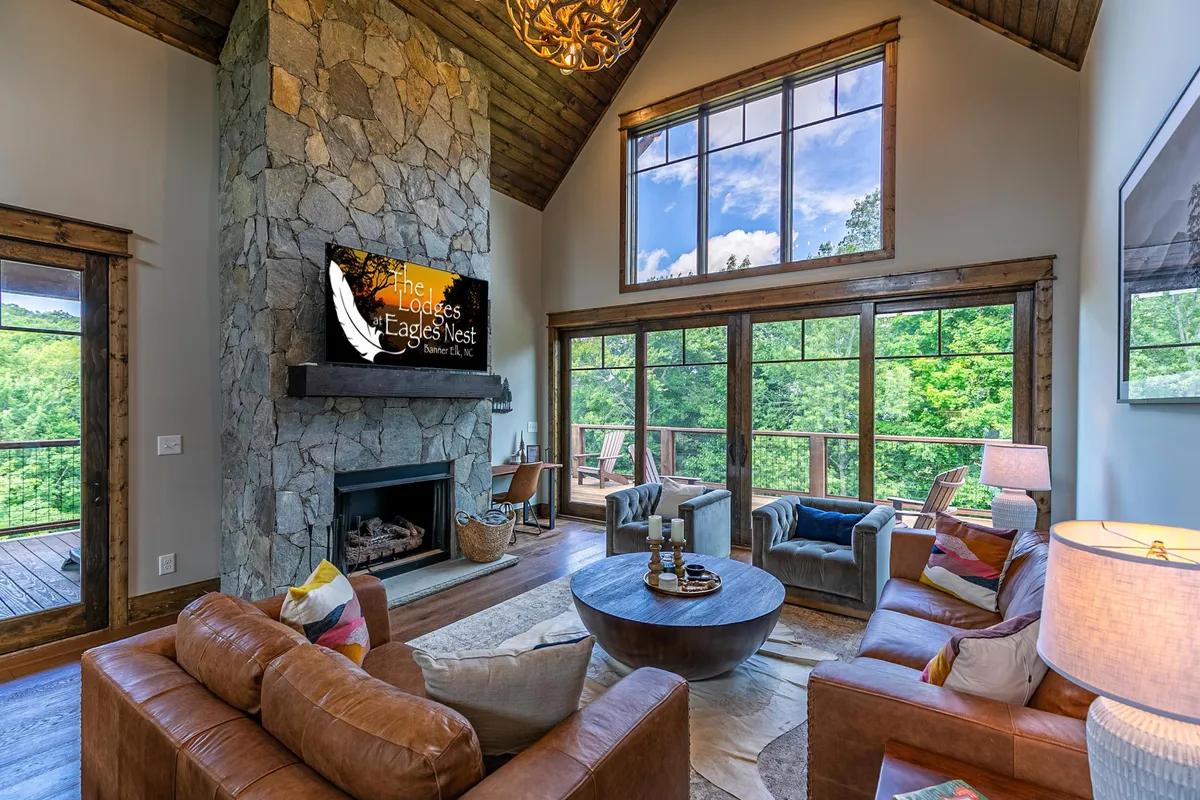 The image presents a welcoming living area featuring a large stone fireplace, an expansive wall of windows with sweeping mountain views, a cozy seating arrangement, and elegant decorative lighting. The television and stylish furnishings add a modern yet homey touch.