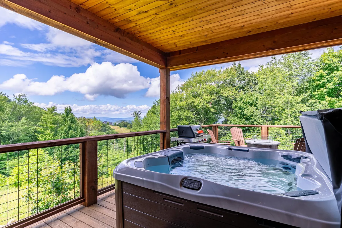 A spacious outdoor deck featuring a private hot tub positioned to maximize sweeping views of the surrounding landscape. The deck is accented by a wooden railing, a grill for barbecuing, and seating areas ideal for conversation or soaking in the scenery. The atmosphere is tranquil and inviting, framed by the natural beauty of the mountains beyond.