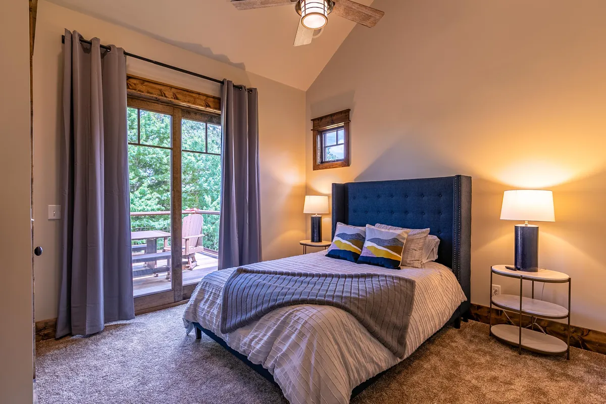 A cozy bedroom with a blue headboard, soft bedding, and a window framing views of the outdoors. A bedside lamp on the nightstand adds to the room's inviting feel, ideal for relaxation or enjoying a morning coffee in solitude.