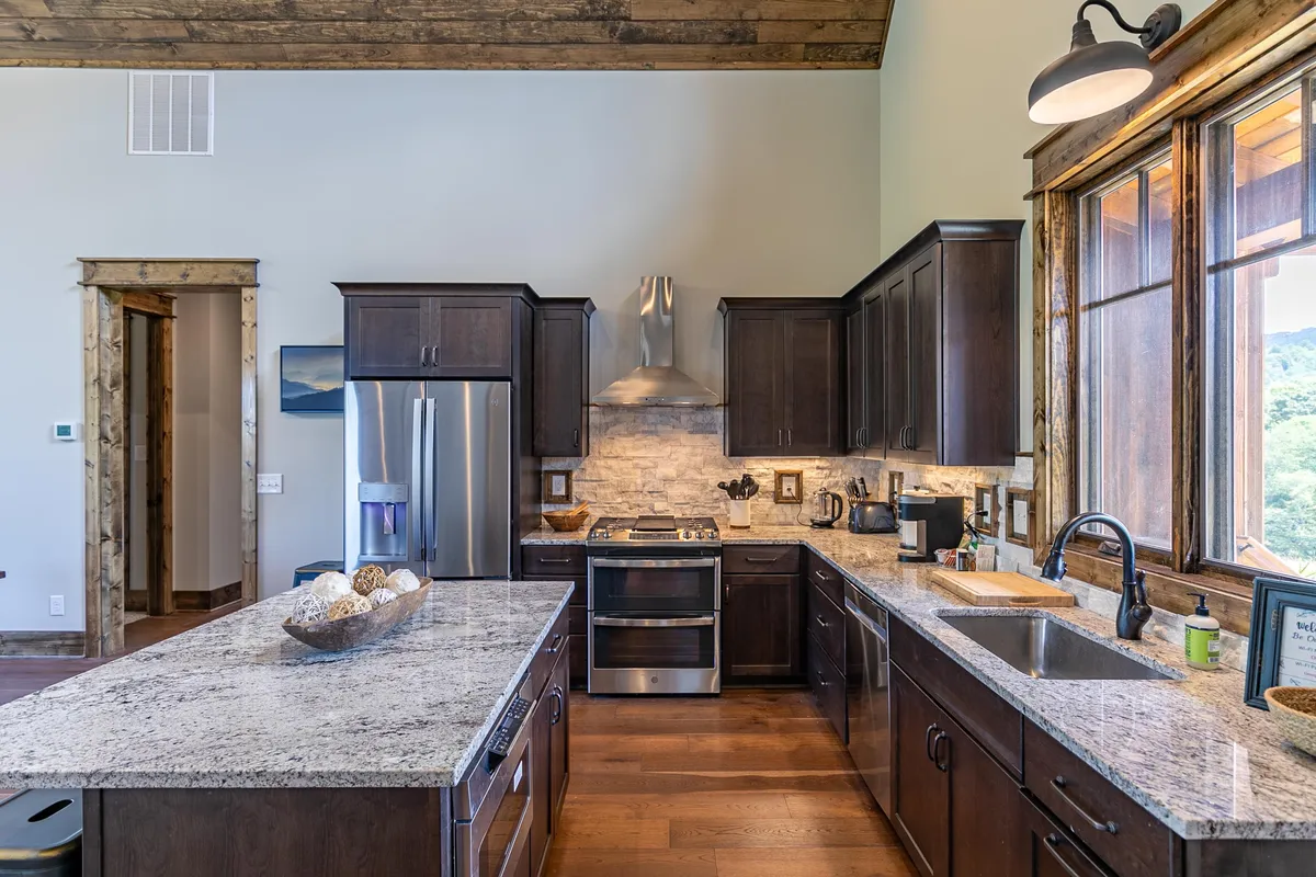 A bright, modern kitchen featuring a granite countertop, sleek stainless steel appliances, wooden cabinets, and expansive windows that flood the space with natural light. The windows offer glimpses of the surrounding landscape, reinforcing the connection between culinary activity and the great outdoors.