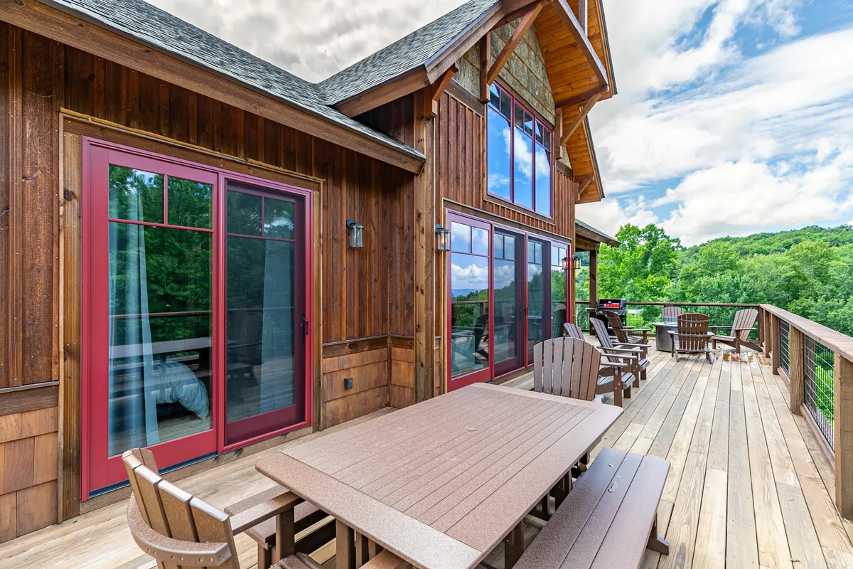 A spacious wooden deck with multiple seating areas overlooks the surrounding natural landscape. The large windows connect the interior to the outdoors, and the deck is framed by trees and greenery, creating an inviting space for relaxation or play with pets.